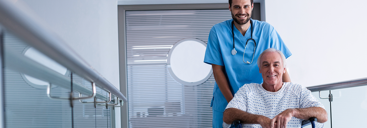 Smiling healthcare worker standing behind an older male patient in a wheelchair in a hospital corridor