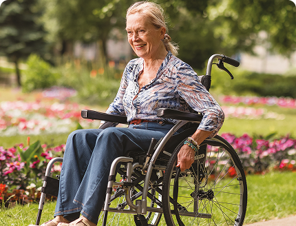 Smiling older woman sitting in a wheelchair outdoors in a flower-filled park.