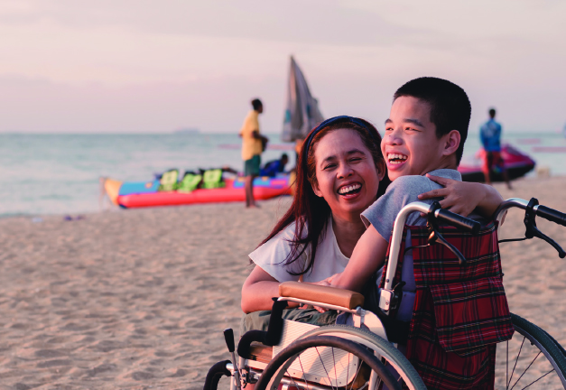 Smiling woman hugging a laughing young boy in a wheelchair on a beach at sunset