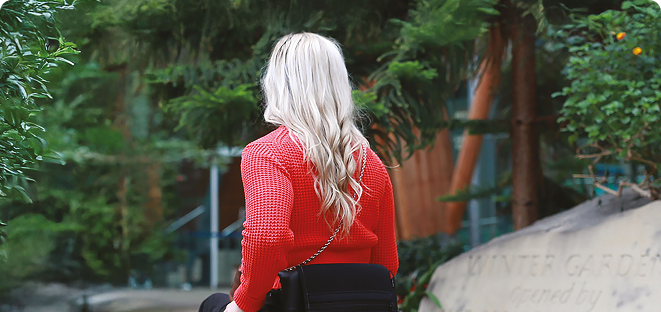Woman with long blonde hair in a red sweater sitting in a wheelchair outdoors near greenery