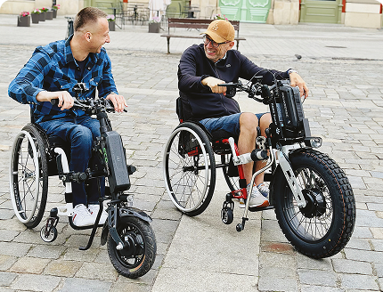 Two men in wheelchairs with electric handbike attachments talking and smiling outdoors