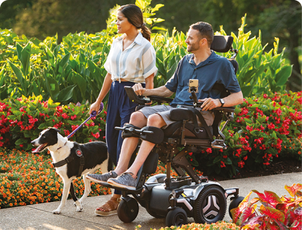 Woman walking a service dog beside a man using a powered wheelchair in a garden
