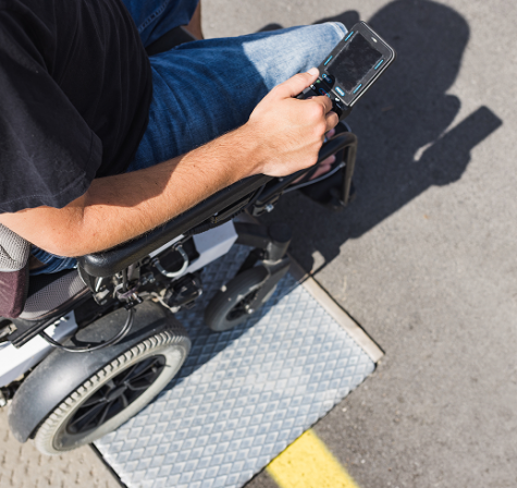 Overhead view of a man sitting in a wheelchair outdoors, holding and looking at a handheld device, positioned on a metal access ramp next to a paved surface.