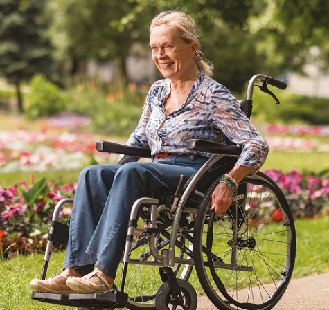 Smiling older woman sitting in a wheelchair outdoors in a flower-filled park.