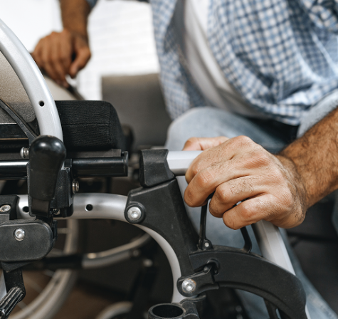 Close-up of a man’s hands adjusting or assembling part of a wheelchair, focusing on the frame and armrest mechanism while he sits indoors.