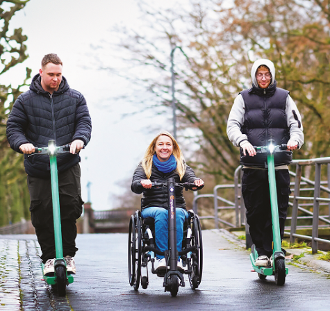 Two men riding electric scooters alongside a smiling woman in a wheelchair with an electric attachment on a path