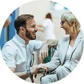 Smiling healthcare worker talking to an older woman sitting in a wheelchair in a hospital hallway